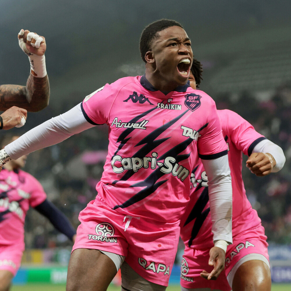 An intense moment as a rugby player in a bright pink jersey shouts triumphantly, surrounded by teammates in a stadium.
