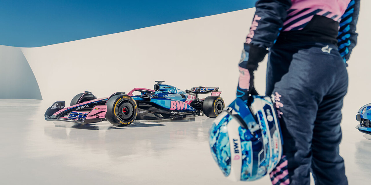 A Formula 1 race car in blue and pink BWT livery stands on a sleek, minimalist set. In the foreground, a race driver in matching suit holds a helmet.