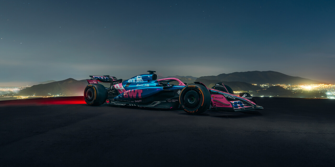 A sleek Formula 1 race car featuring dynamic blue and pink livery is driving on an road at night.