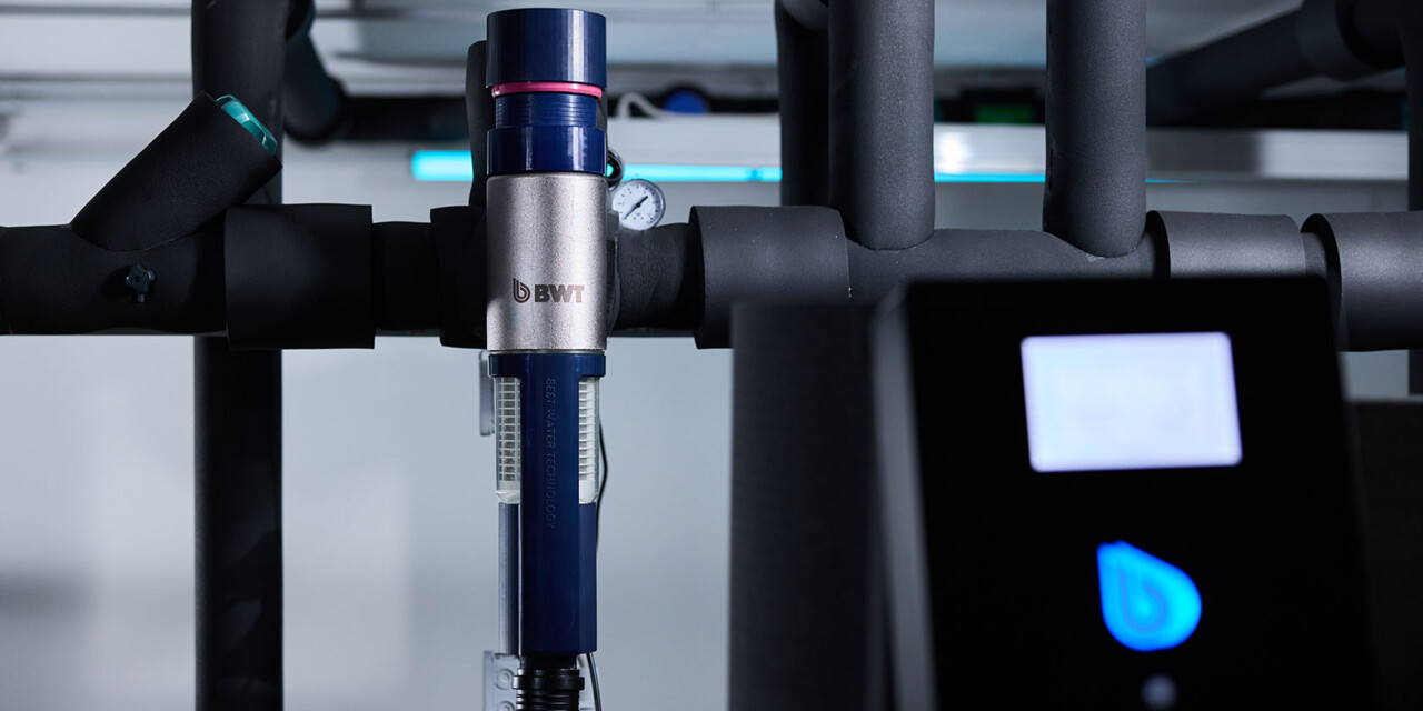 Close-up view of a modern BWT-branded water filtration unit integrated into a network of black pipes. 