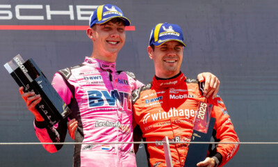 Two professional race car drivers stand on the podium celebrating victory at a Porsche motorsport event.