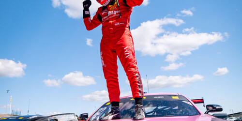 A professional race car driver stands triumphantly atop a pink BWT race car under a bright blue sky dotted with clouds.
