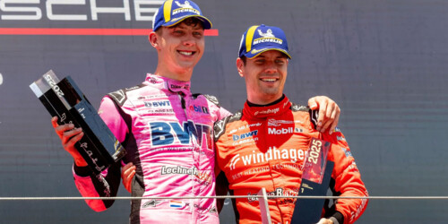 Two professional race car drivers stand on the podium celebrating victory at a Porsche motorsport event.