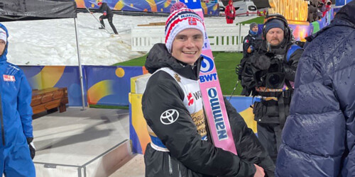 A smiling ski jumper poses with brightly pink colored skis at a professional winter sports event.