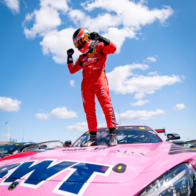A professional race car driver stands triumphantly atop a pink BWT race car under a bright blue sky dotted with clouds.