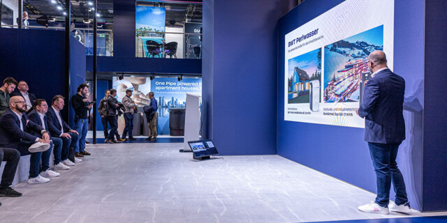 A professional presenter delivers a business presentation to an attentive audience at a modern trade show booth.