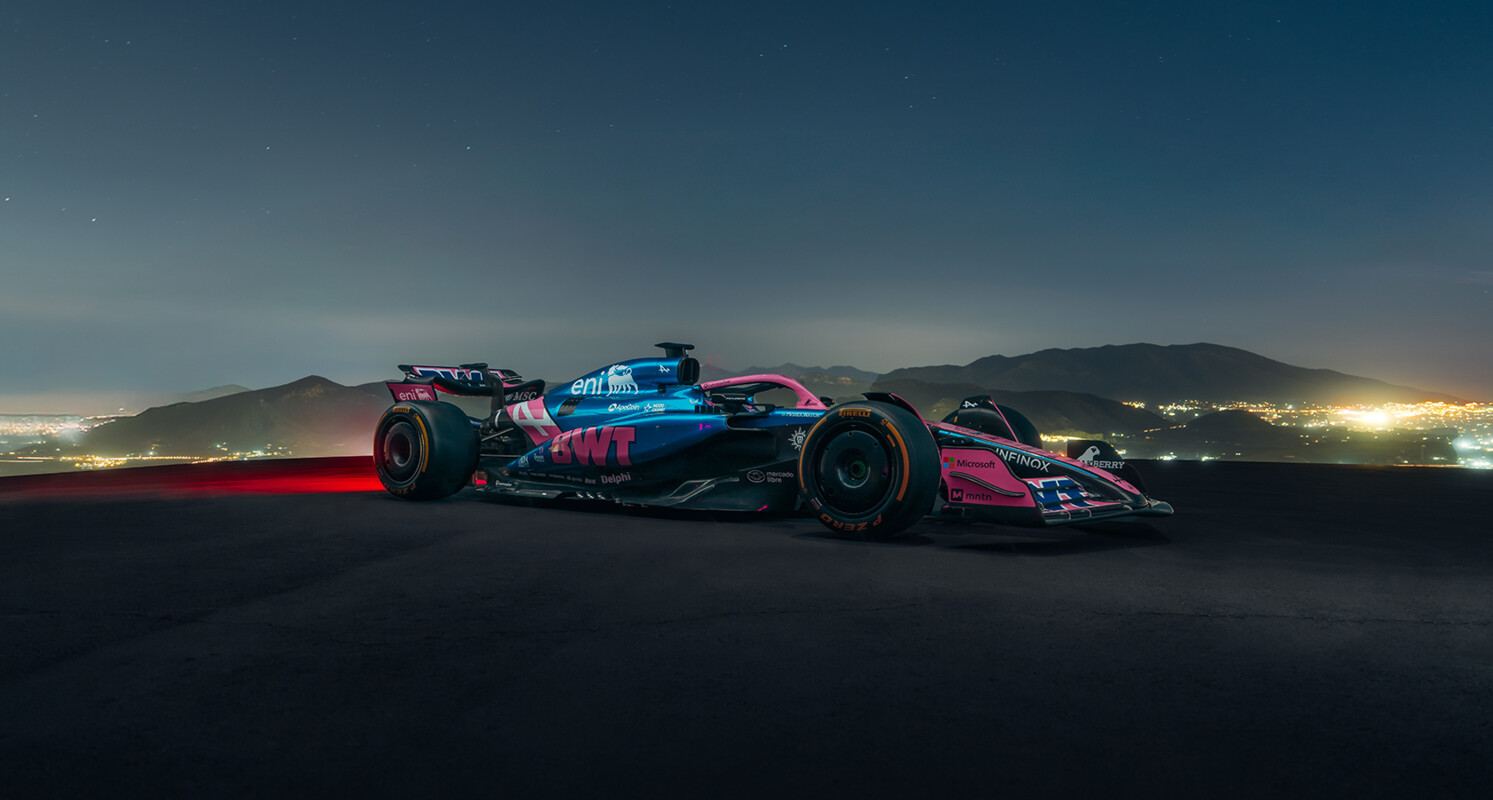 A sleek Formula 1 race car featuring dynamic blue and pink livery is driving on an road at night.